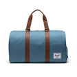 A blue duffel bag with brown leather straps and a small Herschel logo patch.
