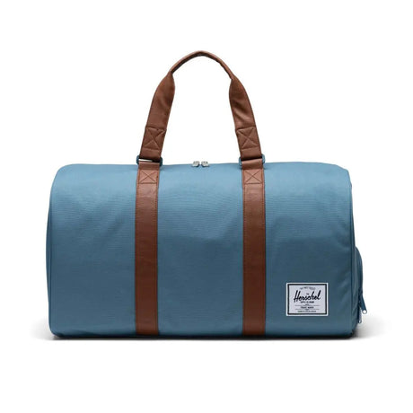 A blue duffel bag with brown leather straps and a small Herschel logo patch.