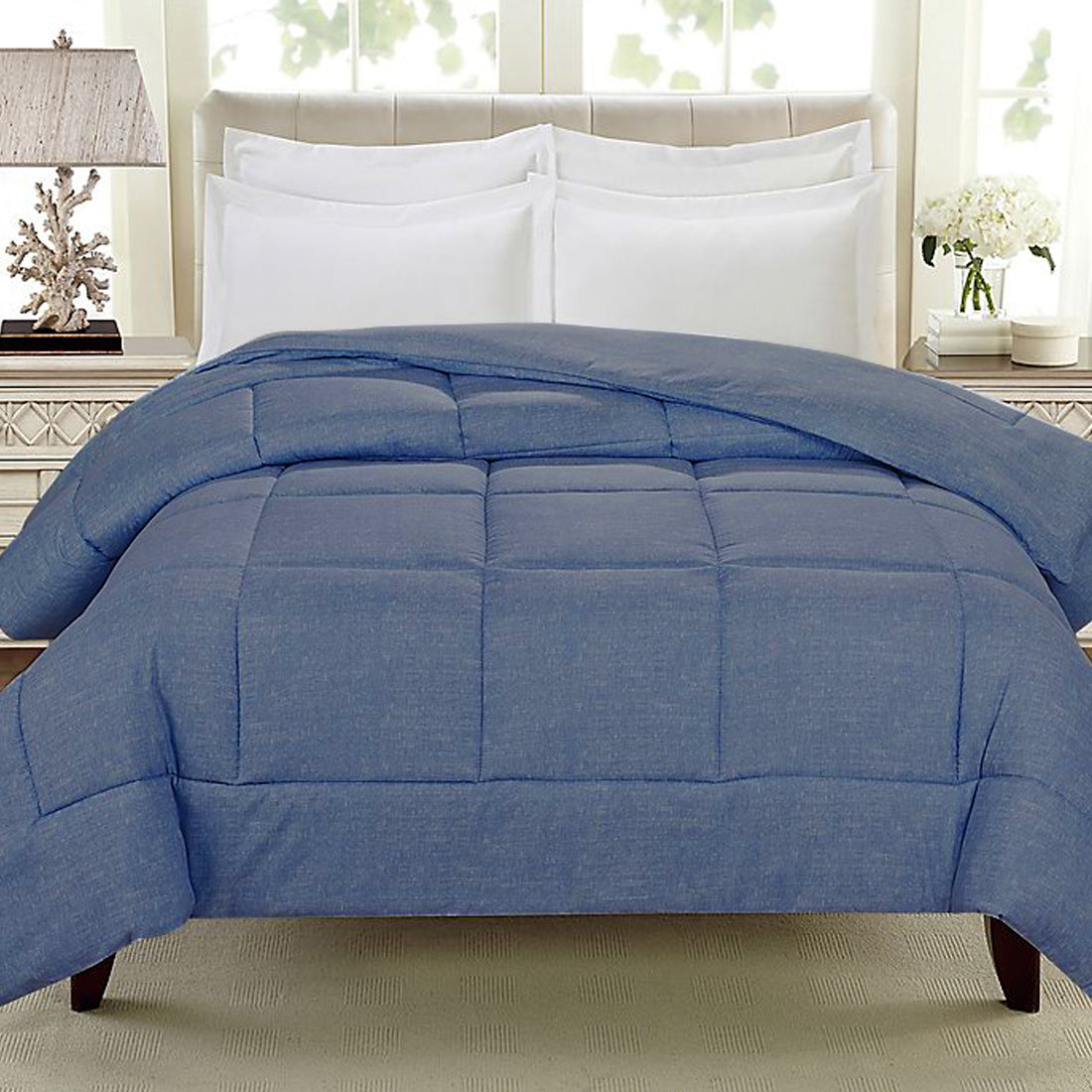 A neatly made bed with a blue quilt and white pillows against a light-colored headboard, with a bedside lamp and decorative elements in the background.