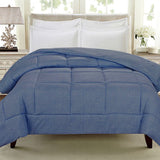 A neatly made bed with a blue quilt and white pillows against a light-colored headboard, with a bedside lamp and decorative elements in the background.
