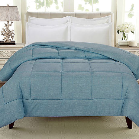 A blue comforter is draped over a bed with white pillows and a light-colored headboard, in a well-lit room with a lamp and decorative items on a nightstand.