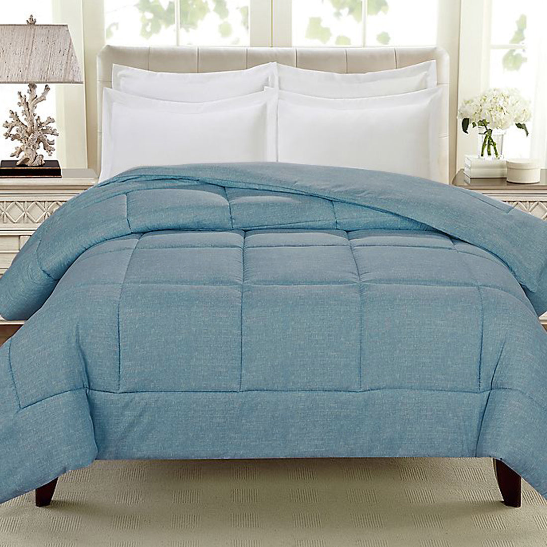 A blue comforter is draped over a bed with white pillows and a light-colored headboard, in a well-lit room with a lamp and decorative items on a nightstand.
