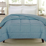 A blue comforter is draped over a bed with white pillows and a light-colored headboard, in a well-lit room with a lamp and decorative items on a nightstand.